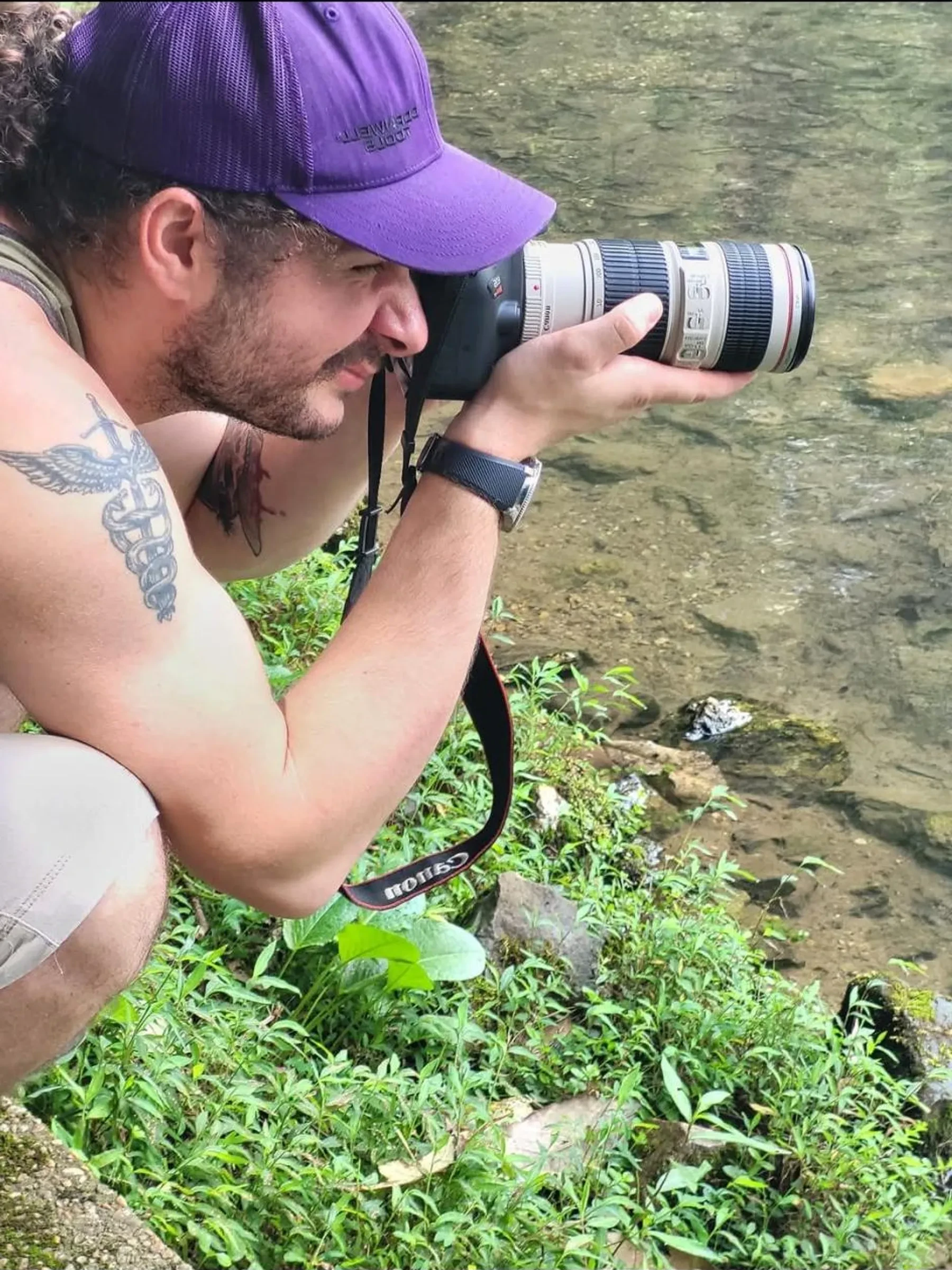 A photographer in a purple cap using a camera with a long lens beside a creek.